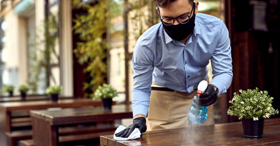 restaurant employee wearing mask cleaning table with cleaner and paper towel