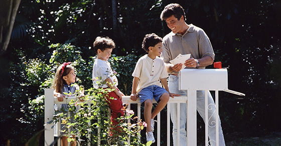 father with two boys and one girl checking mail. Boys are sitting on white fence while dad holds mail.