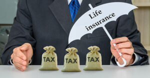 close up of businessman sitting at a table holding a paper umbrella that says life insurance over 3 small burlap sacks on the table that are each labeled TAX