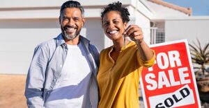 couple standing in front of home with sold sign holding out keys smiling