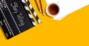 yellow background with movie scene clapboard with title "storytelling".  Sharpened pencils, paper and coffee cups next to clapboard.