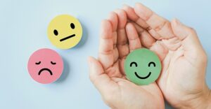 pink sad face icon and yellow indifferent face icon on light blue background next to a pair of hands holding a green smiling face icon