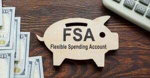 flat wooden piggy bank with letters FSA and then Flexible Spending Account written under FSA.  Calculator and money in miscellaneous denominations on either side of piggy bank