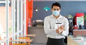 server standing in empty cafe giving a thumbs up sign while wearing mask and holding clipboard