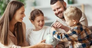 mother, father and two kids smiling around piggy bank as dad teaches son how to put money in