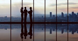 silhouette of 3 business people against floor to ceiling windows with a city skyline in the back
