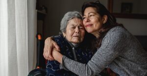 Elderly mother sitting in chair while daughter sits on arm of chair with arms around mother in an embrace.