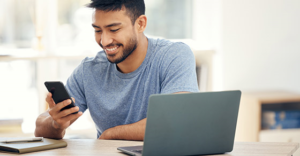man sitting at laptop while looking at phone and smiling