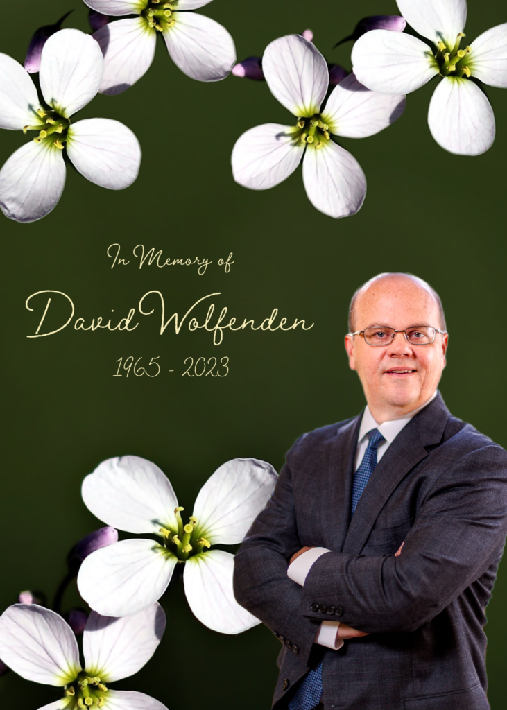 photo of David Wolfenden in suit standing smiling at camera with arms crossed in front of him. Background is dark green, almost black with large forget me not flowers around perimeter and In Memory of David Wolfenden 1956-2023 written in script font
