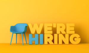 Yellow background with stacked words in yellow stating We're hiring. The HI in hiring is blue and next to the stacked words is a blue chair.