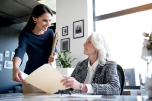 Low angle of cheerful elegant women are looking at each other with smile. Older lady is sitting at table and holding folder while her young boss is standing with documents near desk in office