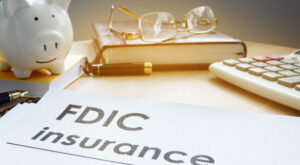 paper that read FDIC insurance surrounded by piggy bank, calculator and glasses sitting on a book.
