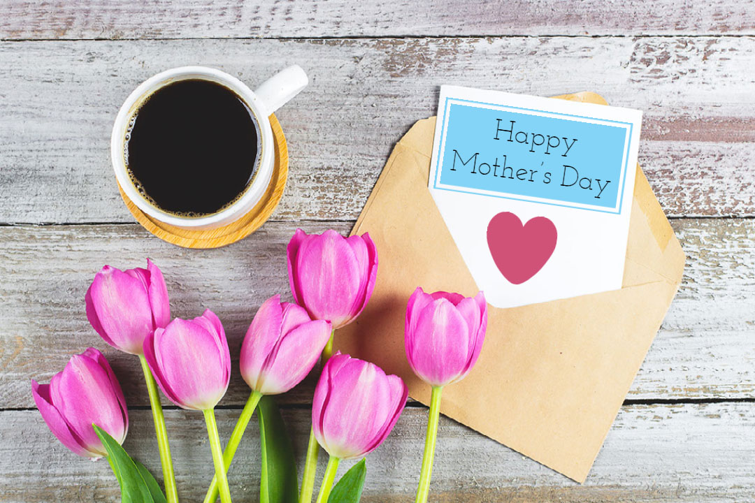 tulips and cup of coffee next to envelope open with happy mother's day and a heart on a sheet of paper sticking out of envelope