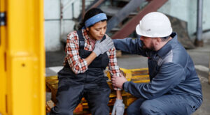 Mature foreman in workwear giving first aid to female subordinate with pain in her shoulder after injury at work in warehouse
