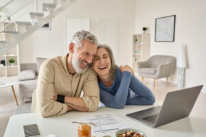 Smiling middle aged senior 50s husband and wife having fun satisfied with buying insurance, paying bills online while enjoying retirement.