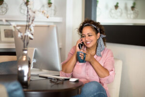 Mixed race woman working from her home office. Sitting smiling at computer while holding a coffee cup in one hand and phone to ear with other hand.
