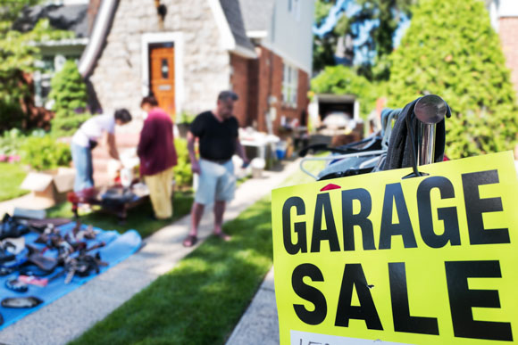 close up of Garage sale sgn with yard sale in the background with unrecognizable people.