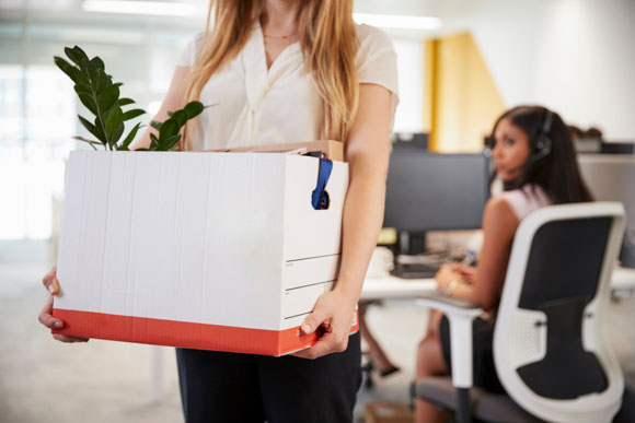 female employee holding box of belongings and walking away from desk after resigning from company while coworker watches in background. 