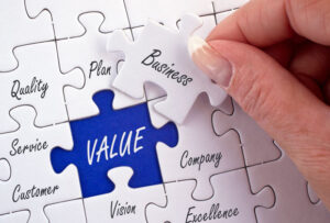 white puzzle with different business terms on each piece. Puzzle piece that says Business on it is being held in a hand and the spot where it fits into the rest of the puzzle is a blue background that says value on it.