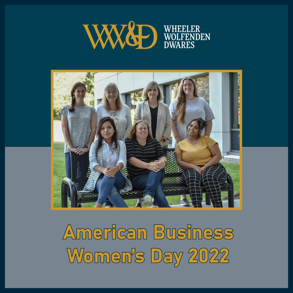 7 women posing on bench in front of office building. Four women standing behind bench and three sitting on bench. Women posing for American Business Women's Day