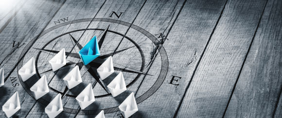 Blue Paper Boat Leading A Fleet Of Small White Boats With Compass Icon On Wooden Table With Sunlight - Strategic Leadership Concept