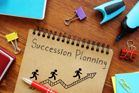 spiral bound notebook with "succession planning" written on the cover. Illustration of figures climbing an upward arrow, surrounded by office supplies such as pens, binder clips, highlighters and folders all on a wooden desk.